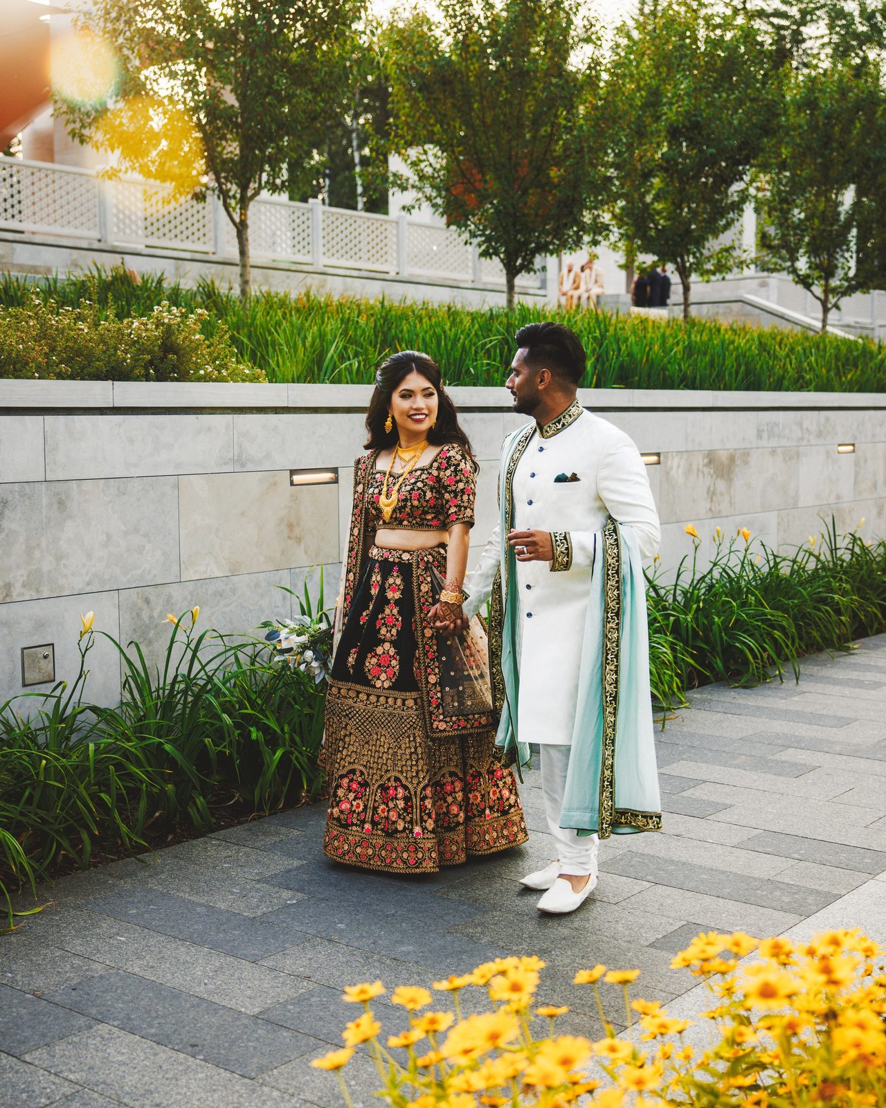 A couple walk a garden path in low evening light, yellow flowers in the foreground.