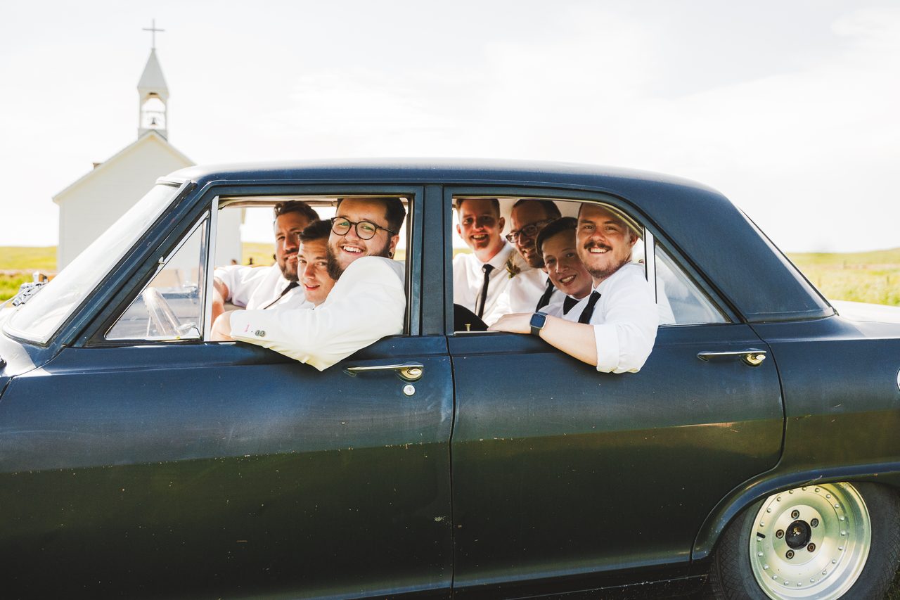The groomsmen crowd out of the windows of a dark vintage car, laughing, a small white prairie church behind them.