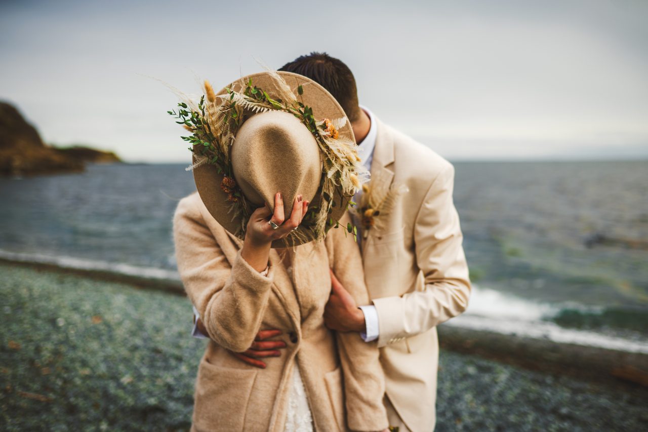 A couple lean into each other on a grey pebble shore, a wide felt hat trimmed with dried flowers held up to hide the kiss.