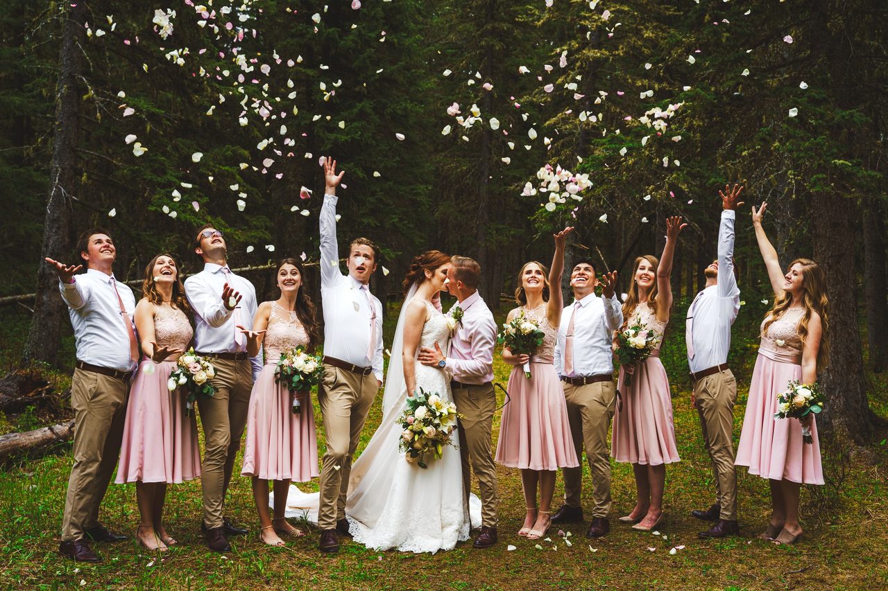 The wedding party throw their arms up together as petals fall under the trees.