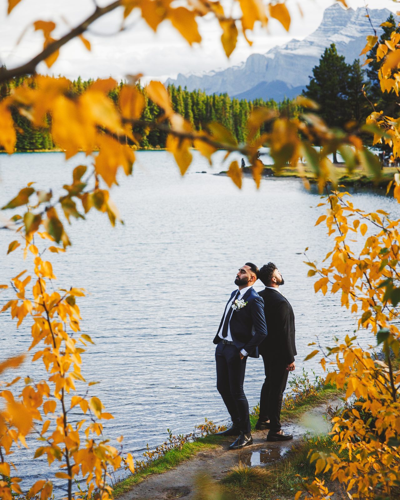 A couple stand by a lake framed by golden autumn leaves.