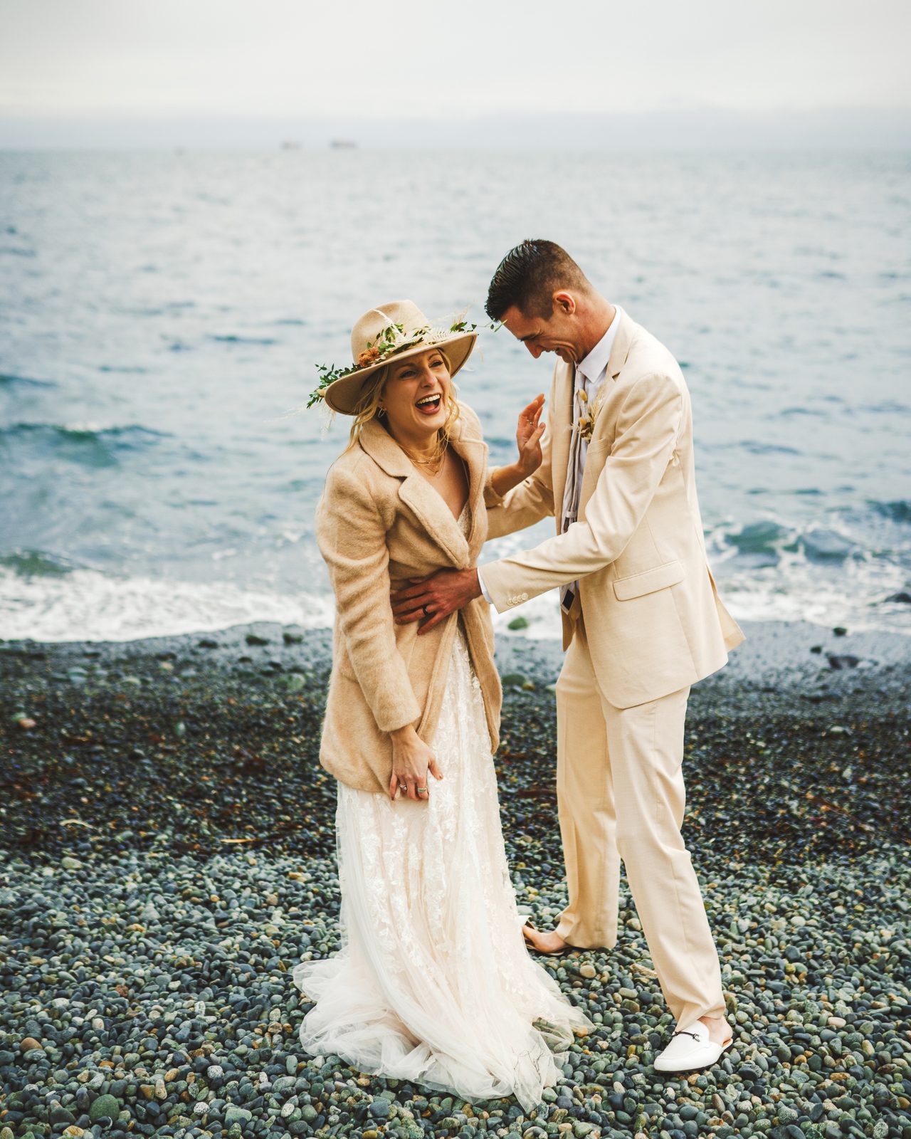 A couple in hats and cream suits stand together on a pebble beach.