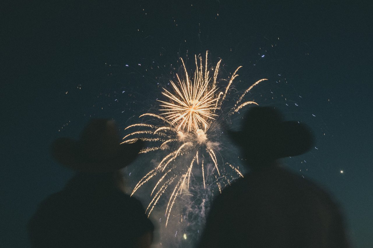 Two figures in cowboy hats watch fireworks burst overhead.