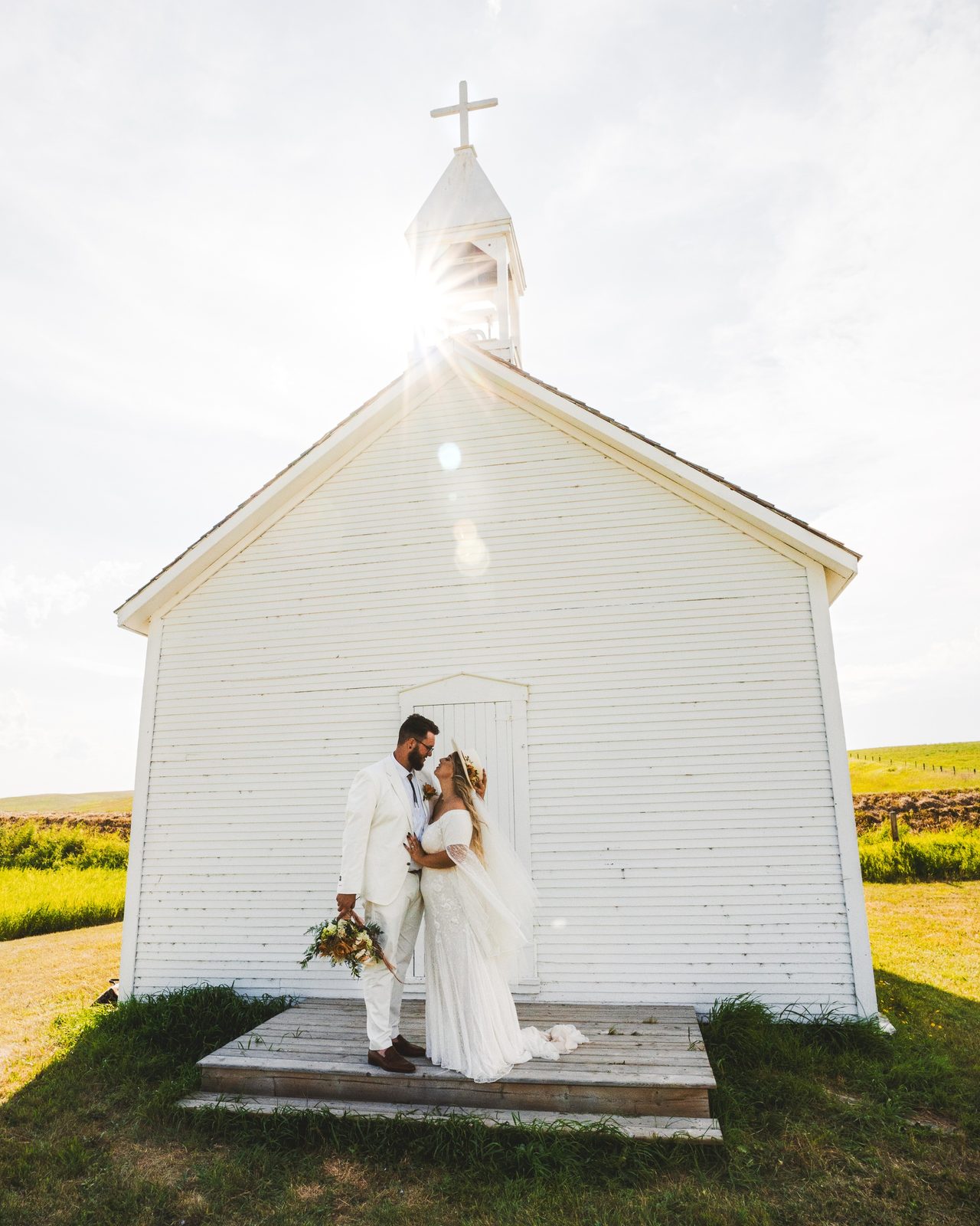 A couple stand before a small white prairie church, the sun flaring behind the steeple.