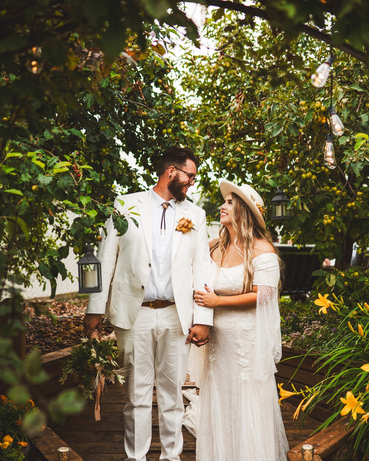 A couple in white stand on wooden steps surrounded by greenery.