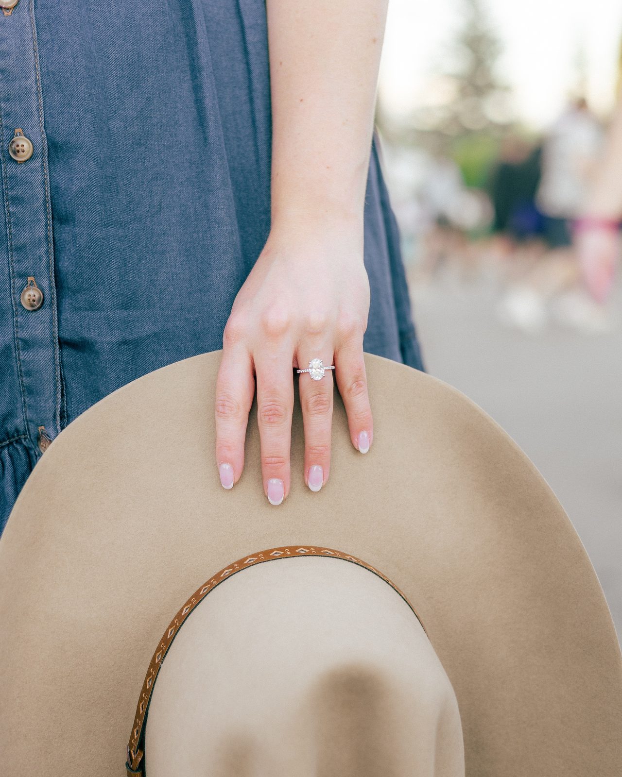 A hand wearing an engagement ring rests on the brim of a felt hat.