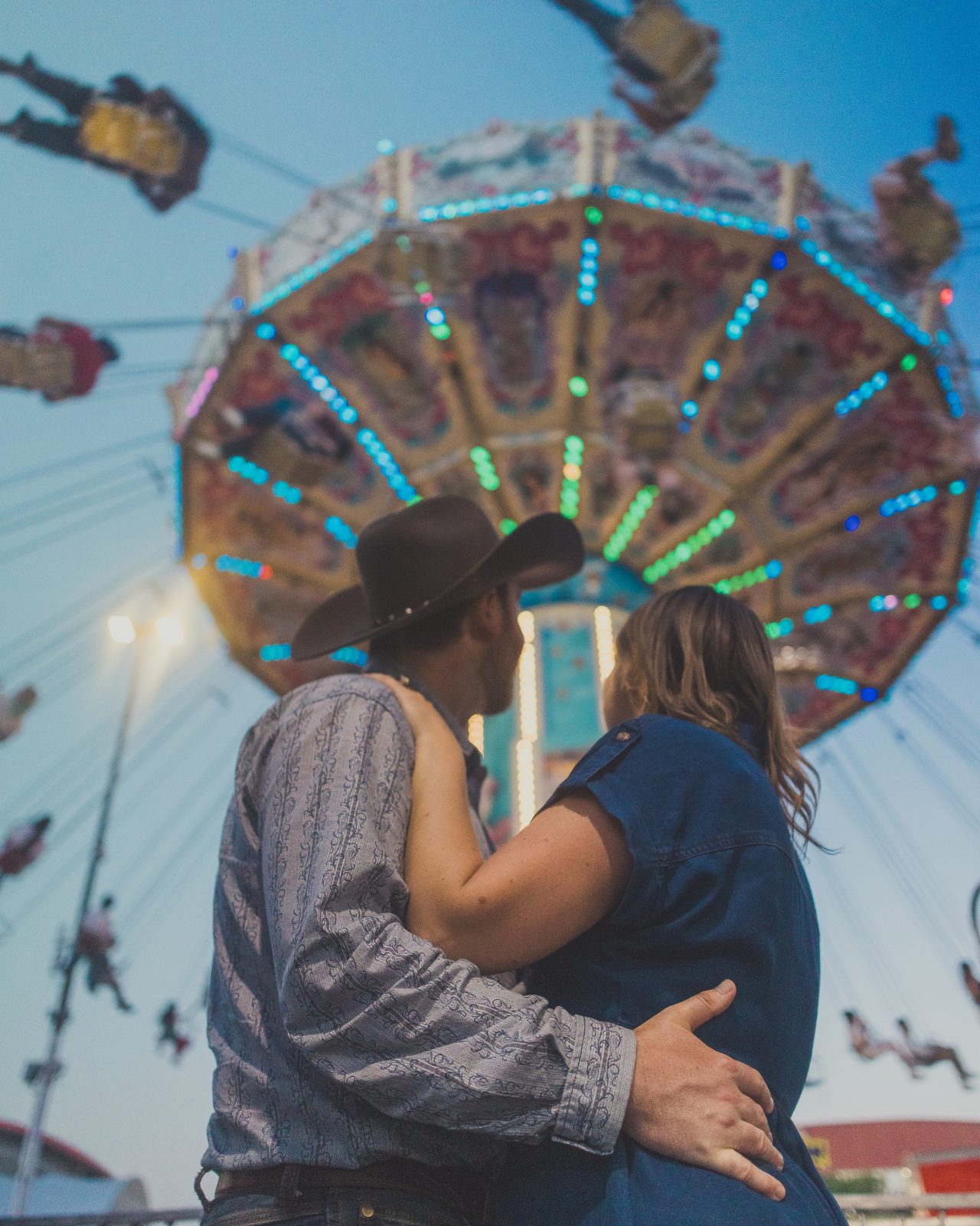 A couple kiss in front of a lit fairground swing ride.