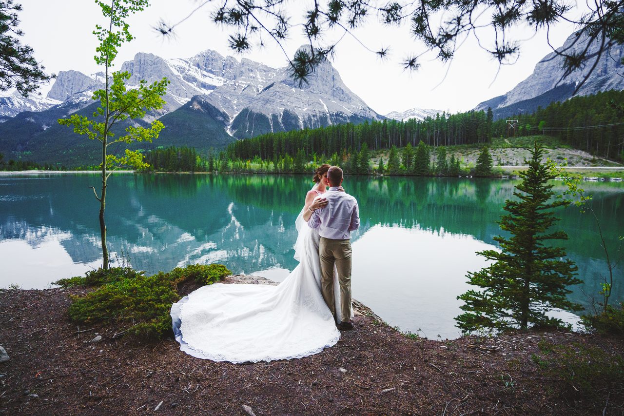 A couple stand at the edge of a turquoise mountain lake, the bride's long train fanned out across the rock, snow still on the peaks behind them.