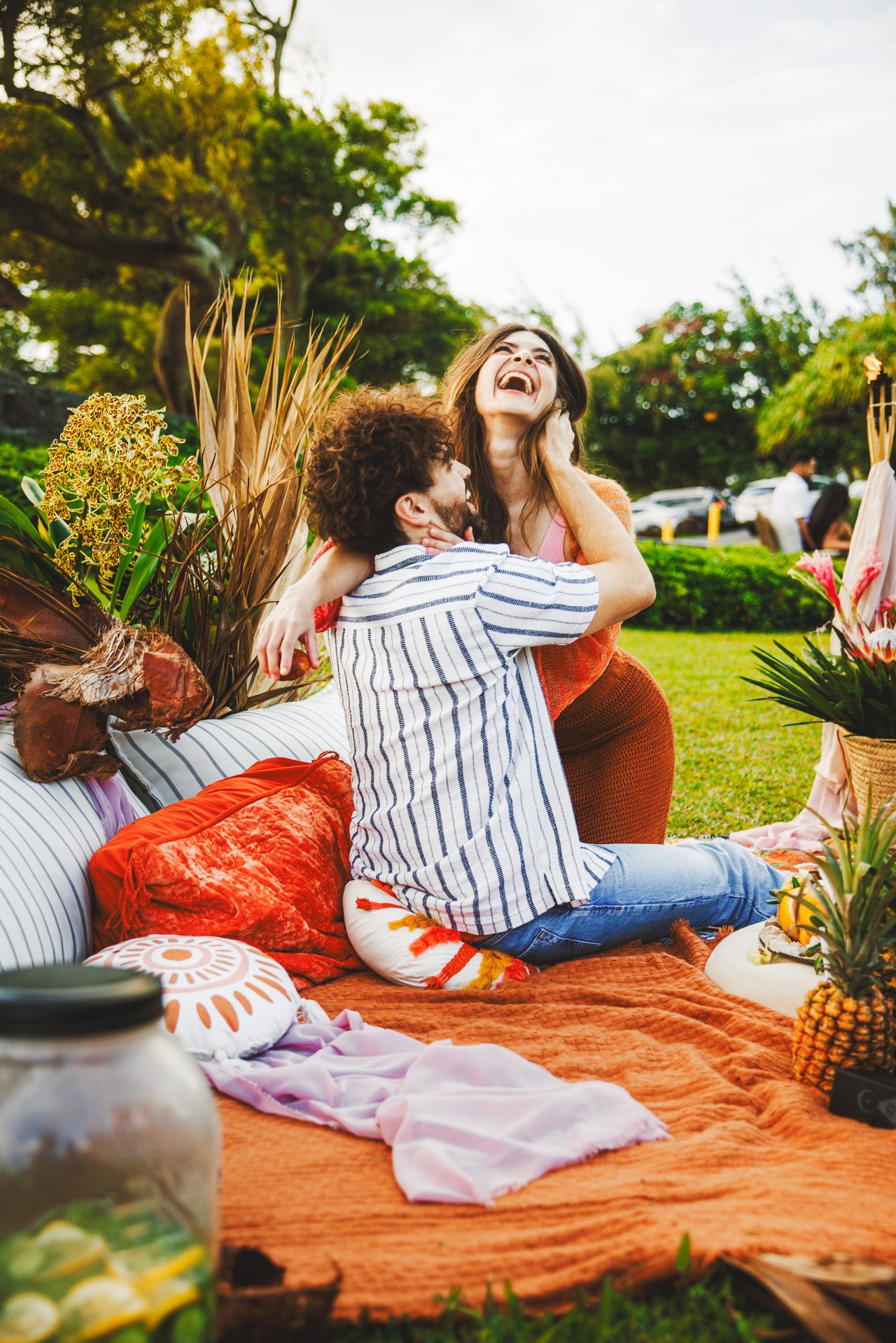 A couple laugh together on an orange picnic blanket, her head tipped right back, cushions and tropical flowers around them.