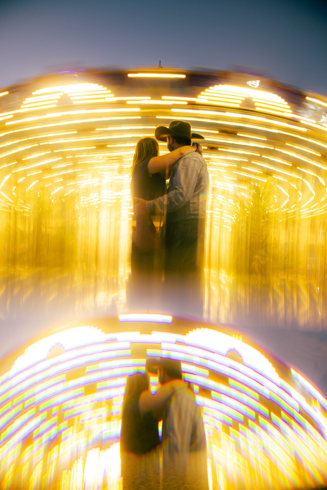 A couple in a cowboy hat hold each other in front of a spinning fairground ride, its lights pulled into long gold streaks around them.