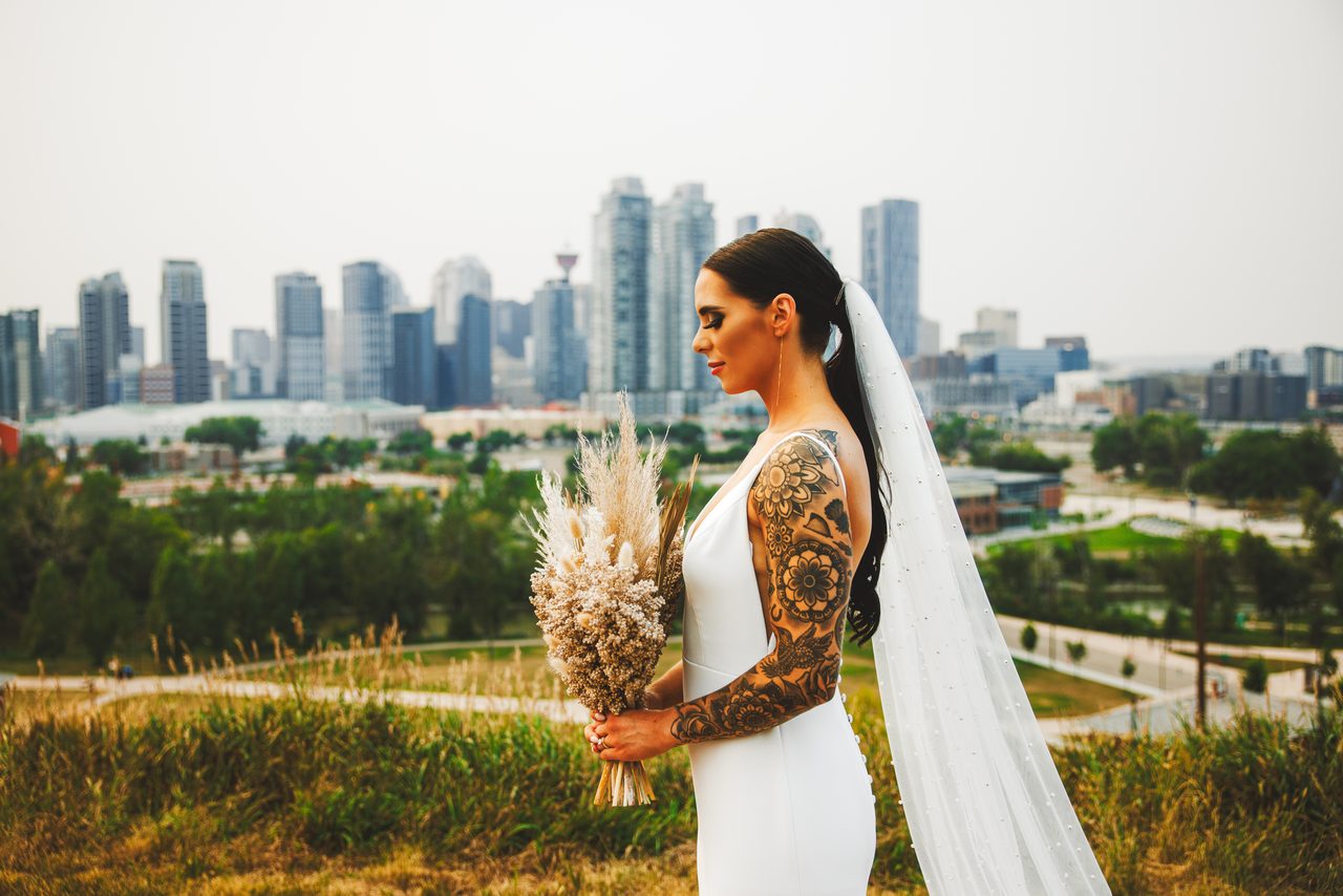 A bride in profile holding a bouquet of dried pampas grass, a floral tattoo down her arm and a long veil, the downtown Calgary skyline behind her.