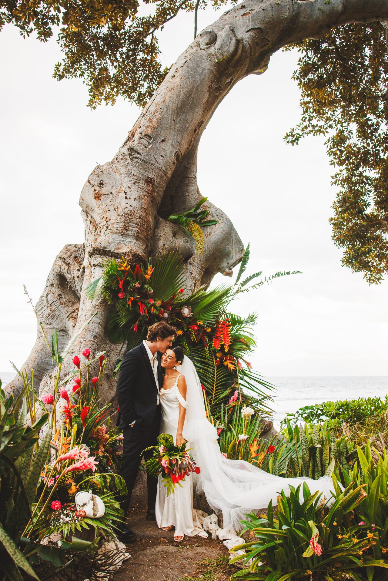 A couple stand close under an enormous leaning fig tree dressed with tropical flowers and palm fronds, the ocean beyond.