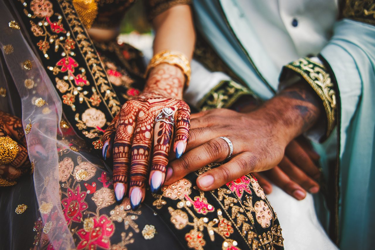 Two hands rest together on an embroidered lehenga — hers patterned with henna and gold bangles, his wearing a wedding ring.