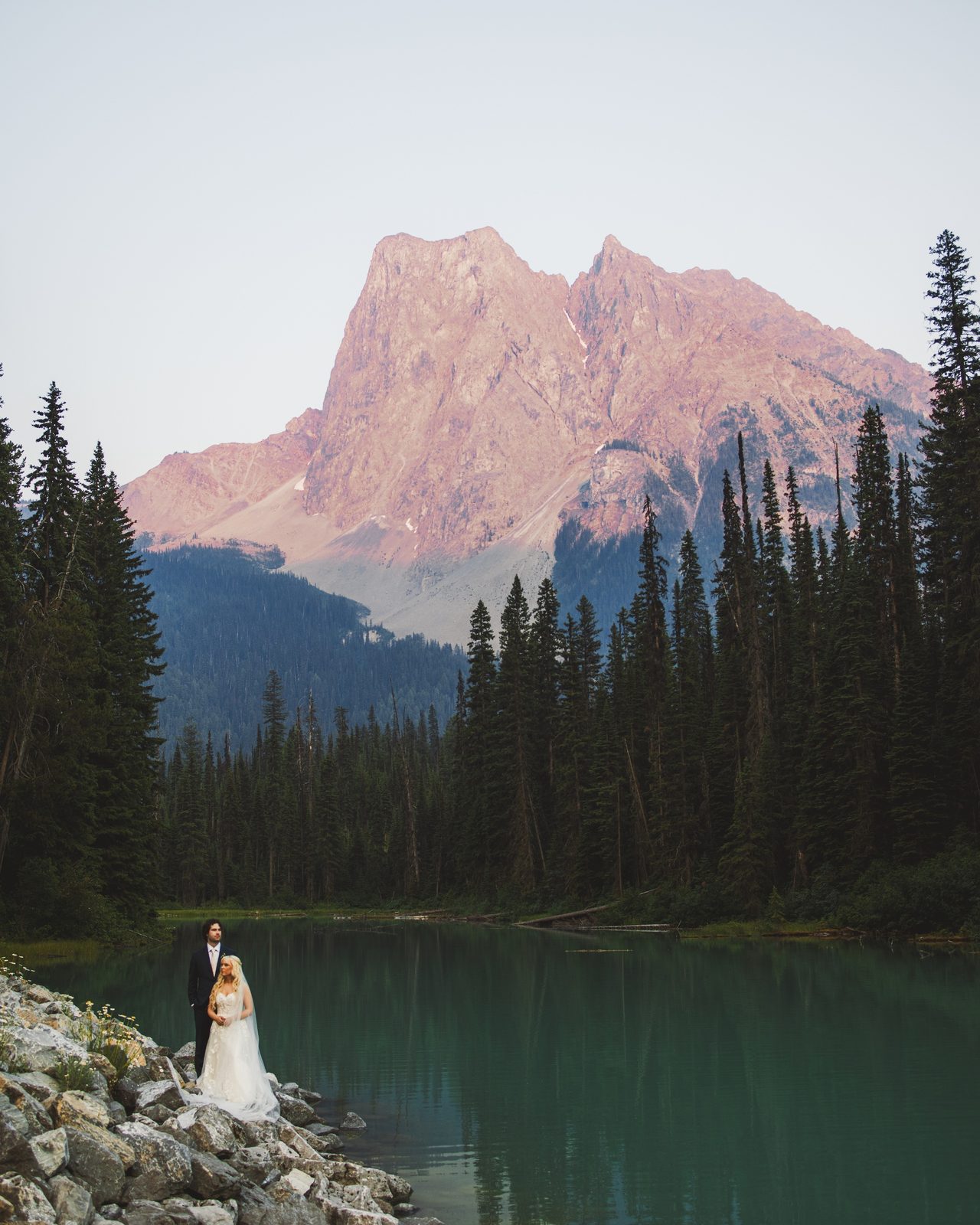 A couple stand at the edge of a mountain lake at dusk on their wedding day.